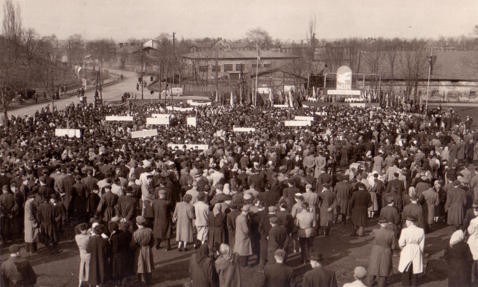 Záběr z manifestace proti zbrojení a nukleárním zbraním v Karviné 14.4.1958. V pozadí, od tribuny vpravo: budova čp. 266 (původně hospodářská budova statku hraběte Larisch-Mönnicha). Uprostřed snímku, za odbočkou se stojícím autem: budova čp. 1023 (za její střechou lze částečně vidět budovu čp. 1314 u železničního nádraží). Vlevo v zatáčce pak budova čp. 67 (Kovárna a zámečnictví Max Herz)