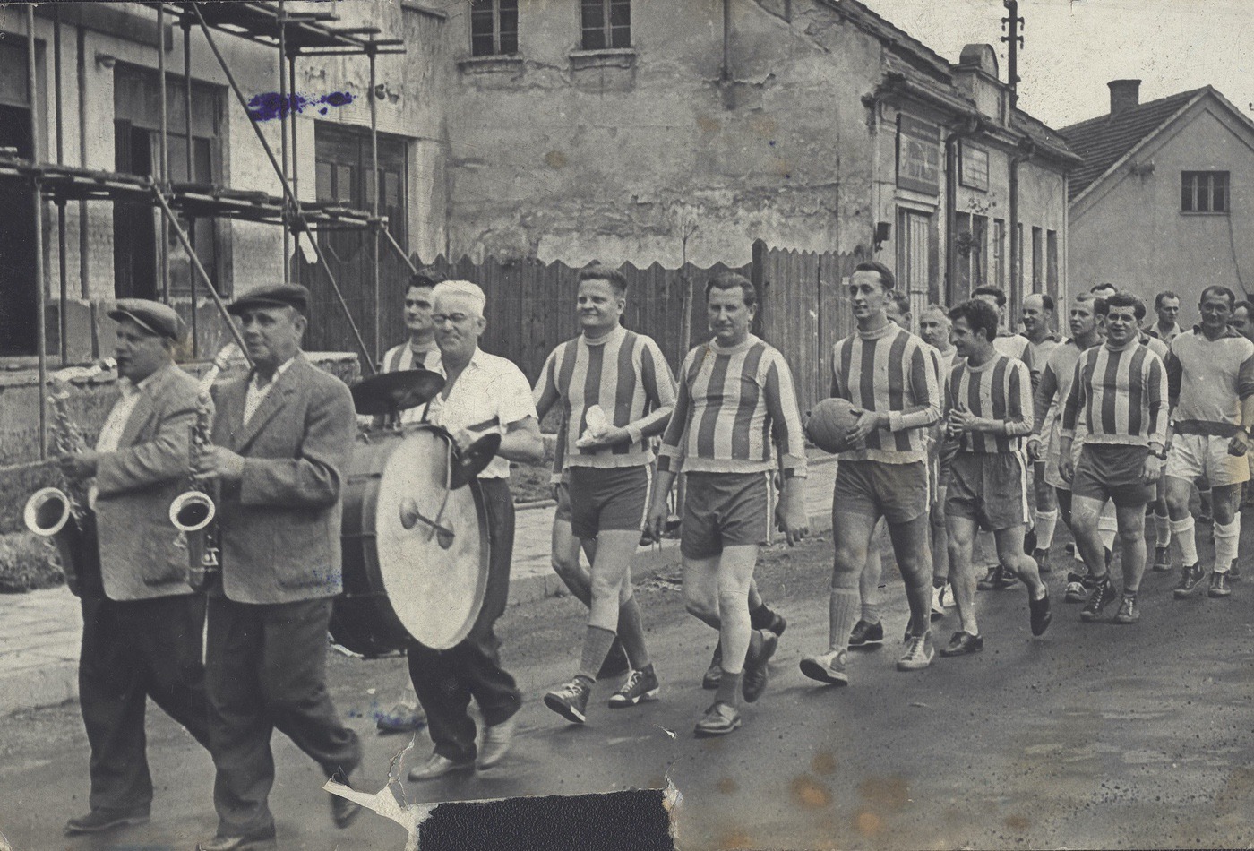 Průvod fotbalistů Slámovou ulicí. Dům s vývěsním štítem: čp. 112 (Potraviny Burysz), vpravo štít domu čp. 24. Fotografováno přibližně v první polovině 50-tých let (fotbalista v bílém dresu úplně vpravo je možná p. František Sochor - autor výstavy a publikace Vzpomínky na starou Karvinnou).